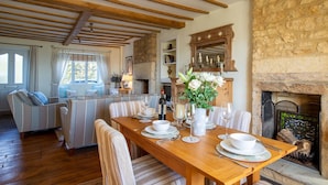 A cozy dining area with a wooden table set for four, surrounded by striped upholstered chairs. A fireplace with a stone wall is visible in the background. - Swift Cottage, Blockley - sleeps 4 guests in 2 bedrooms (Blockley)