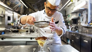 Un chef de cuisine, dans une cuisine professionnelle, tenant une passoire au-dessus d’un plat de dessert.