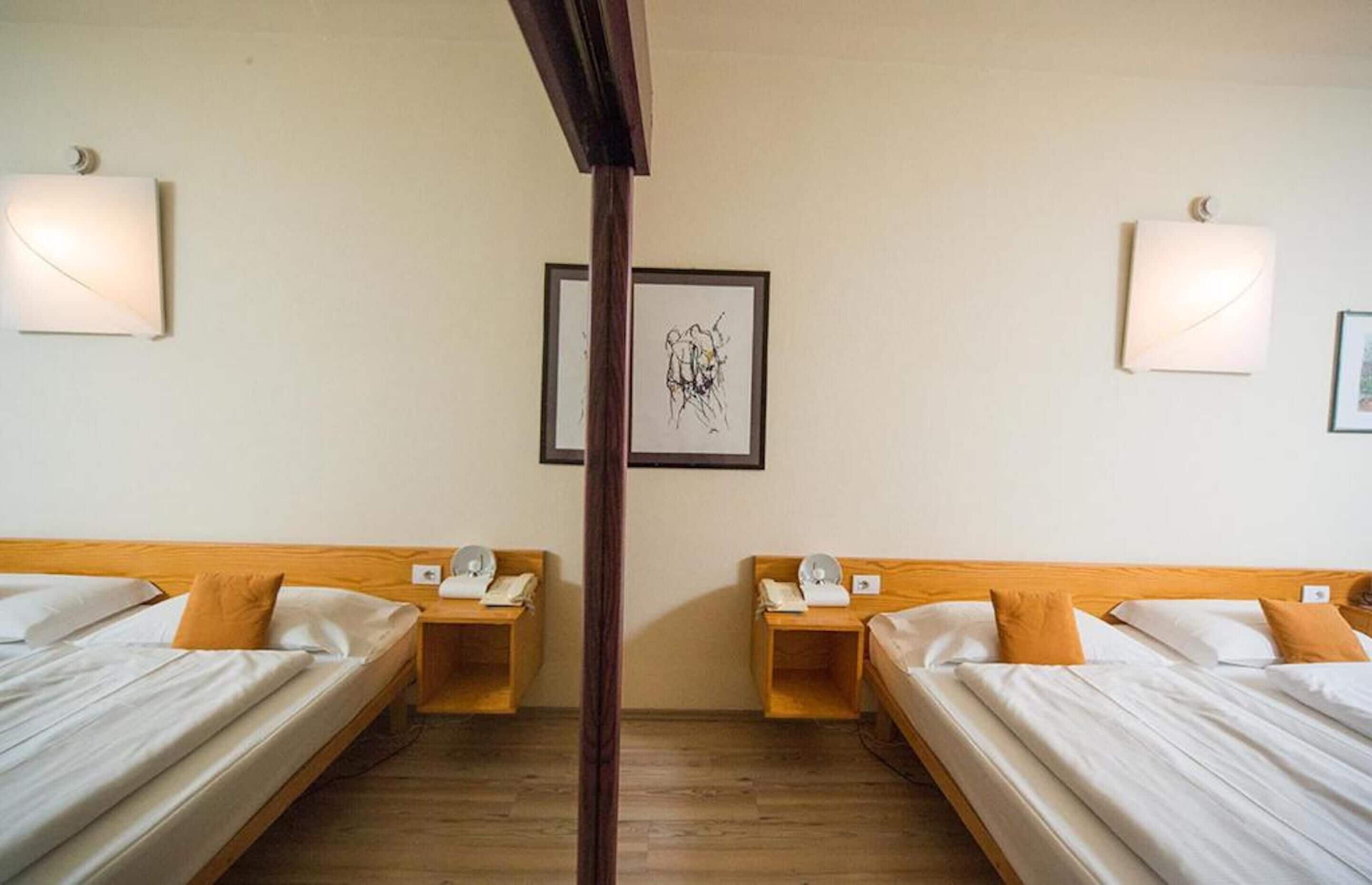 Camera d'albergo con due letti singoli, testiere in legno, comodini con lampade e quadri appesi alla parete.