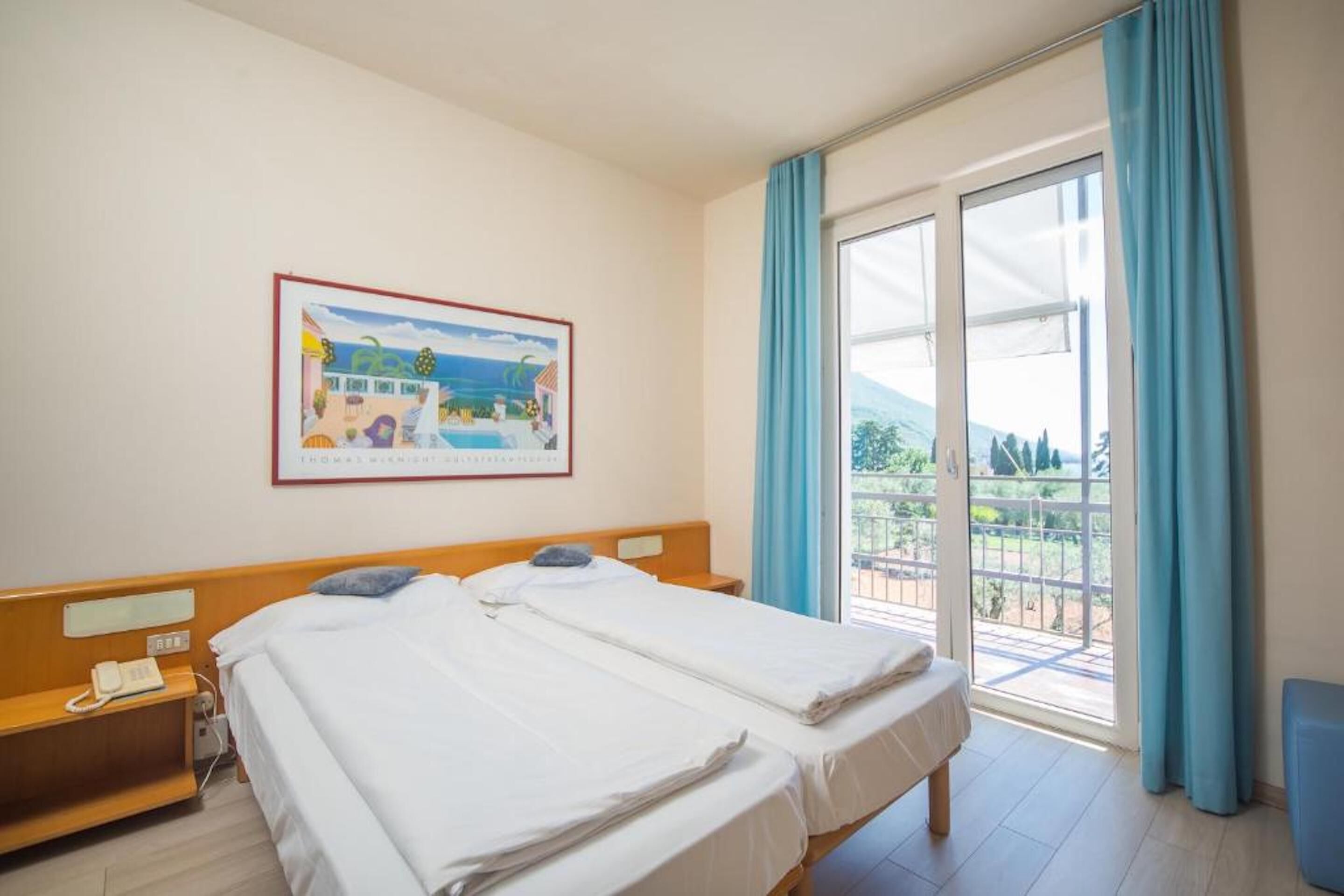 A hotel room with two beds, a wooden headboard, a bedside table with a phone, a painting on the wall, and a balcony with blue curtains.