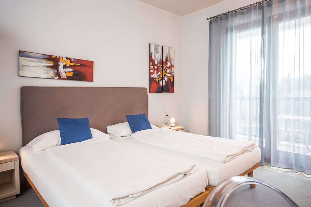 Habitación de hotel con dos camas, una mesita de noche y una ventana con cortinas translúcidas.