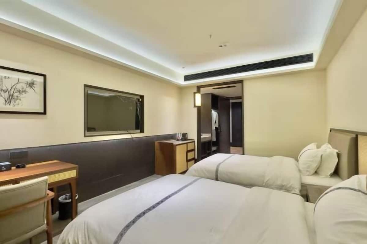 Ein Hotelzimmer mit zwei Betten, einem Schreibtisch, einem Sessel, einer kleinen Kommode, einem Spiegel und einem gerahmten Kunstwerk an der Wand.