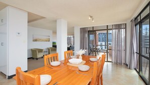 A spacious dining area with a wooden table set for six, surrounded by chairs. A large window offers a view of the cityscape. - Fountain Court 25 - Spacious Unit with Ocean Views (Port Macquarie)