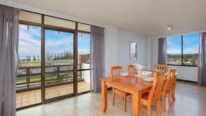 A dining area with a wooden table and chairs, large windows with curtains, and a view of a parking lot and trees. - Fountain Court 25 - Spacious Unit with Ocean Views (Port Macquarie)