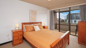 A bedroom with a large bed, wooden nightstands, a desk, and a view through a large window. - Fountain Court 25 - Spacious Unit with Ocean Views (Port Macquarie)