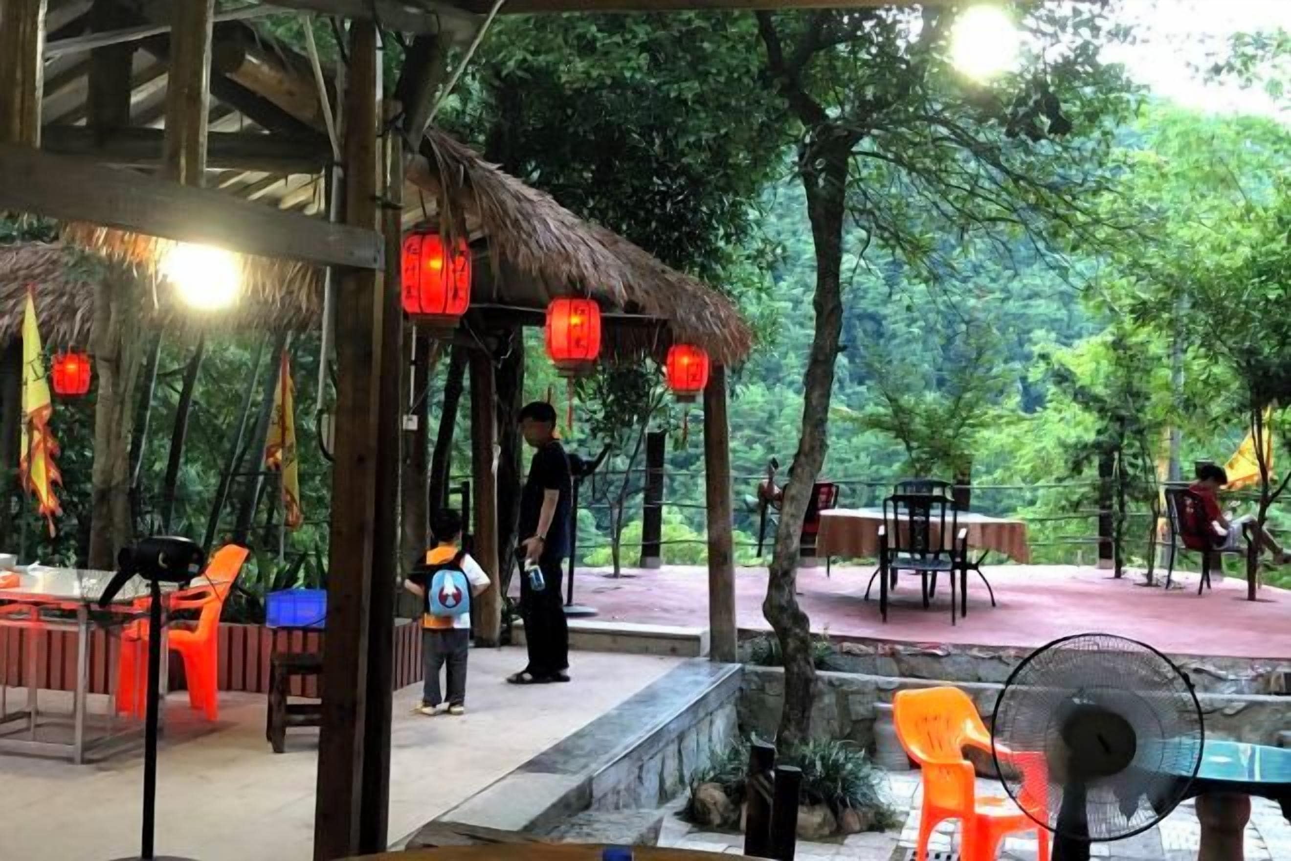 An outdoor dining area with tables and chairs, surrounded by trees and a thatched-roof structure.