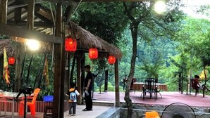 An outdoor dining area with tables and chairs, surrounded by trees and a thatched roof structure. - Hongyezhai Ecological Leisure Villa (Hengyang)