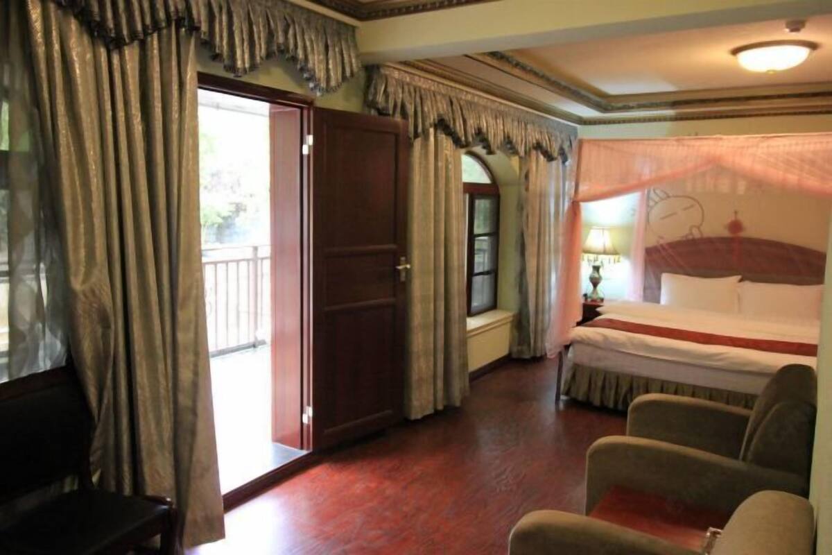 A hotel room with a bed, a bench, a chair, a window with curtains, and a door.