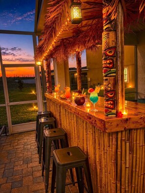 Ein Außenbereich mit Bar, einem Reetdach, Tiki-Dekoration und Blick auf den Sonnenuntergang.