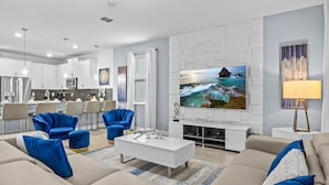 A modern living room with a large TV, blue chairs, a white coffee table, and a kitchen in the background. - Professionally Purpose Built Vacation Oasis- New Discounts Just Applied! (Davenport)