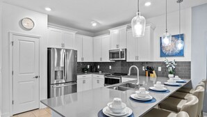 A modern kitchen with white cabinets, stainless steel appliances, a large island with blue placemats, and a hanging light fixture. - Professionally Purpose Built Vacation Oasis- New Discounts Just Applied! (Davenport)