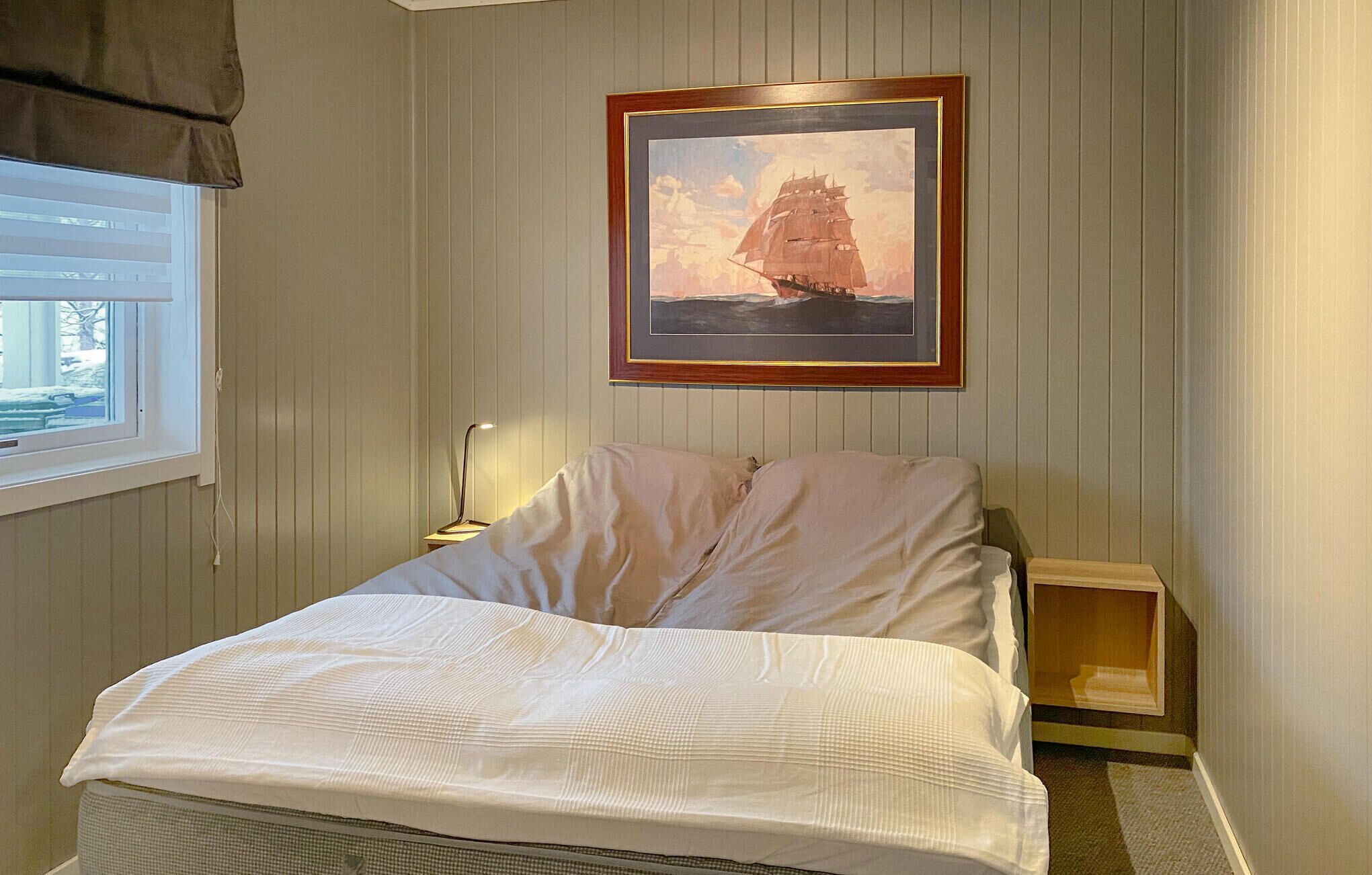 Ein Schlafzimmer mit Bett, Nachttisch, Lampe, Fenster und einem gerahmten Bild an der Wand.