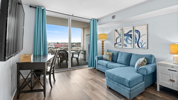 A modern living room with a blue sofa, a dining table with chairs, a TV, and a balcony with a view.