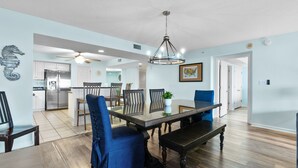 A spacious dining area with a wooden table and blue upholstered chairs, a chandelier, and a kitchen in the background.
