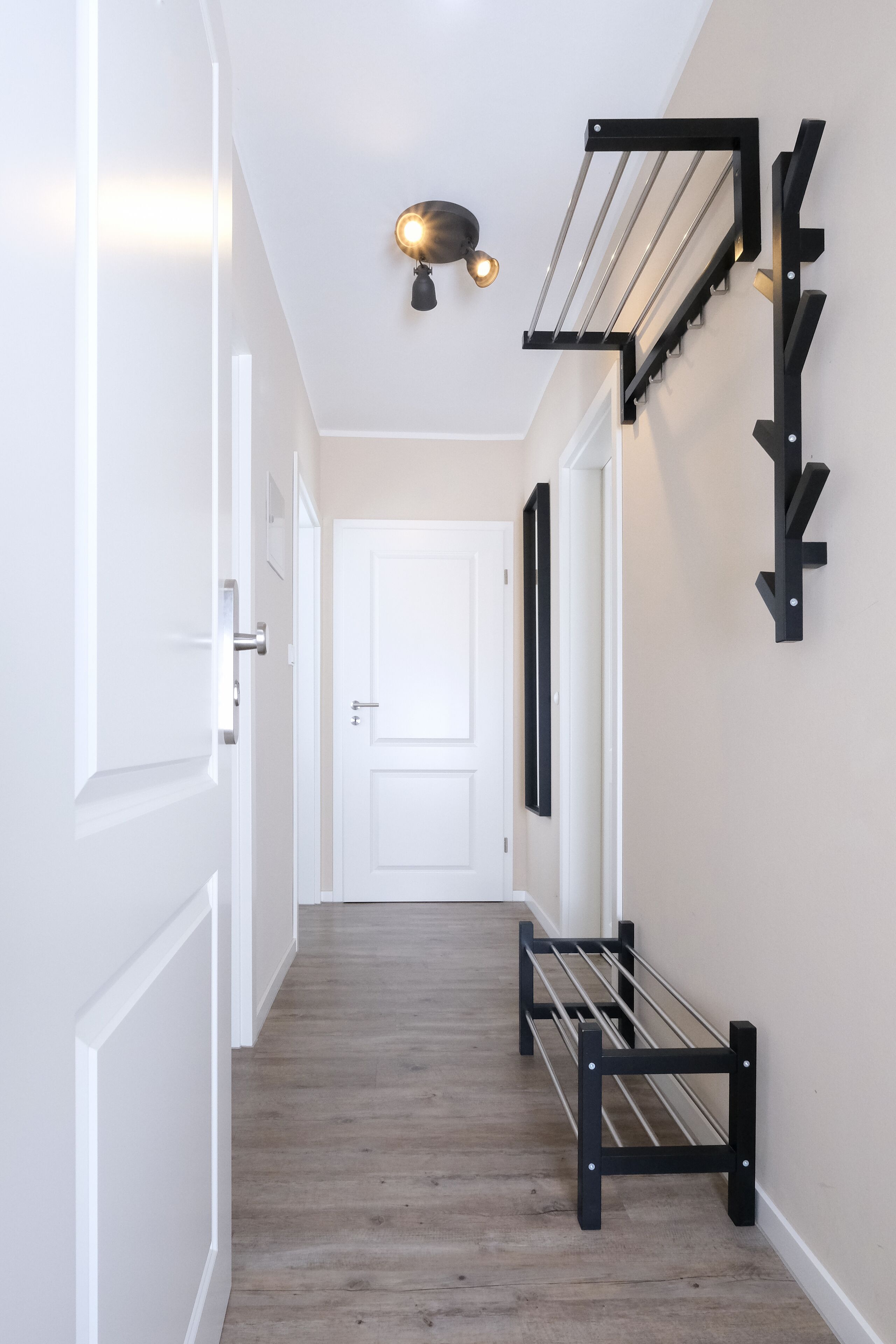 Ein Flur mit Holzboden, weißen Wänden und einer weißen Tür am Ende. An der Wand befinden sich schwarze Metallregale und eine Leuchte darüber.