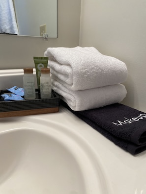 Un lavabo de salle de bain avec une serviette blanche, une serviette noire ornée d’une broderie « Maquillage », et deux flacons de shampoing à la menthe et au romarin de la marque Aveda.