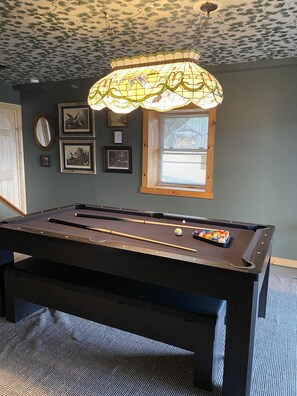 Une table de billard avec un jeu de billard (queue et billes), un luminaire suspendu et des tableaux encadrés au mur.