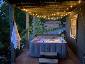 A hot tub under a wooden pergola with string lights, surrounded by greenery and a house in the background.