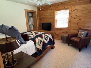 Une chambre avec un mur en bois, un lit recouvert d’une couverture à motifs, une télévision, une chaise et une petite table avec une lampe.
