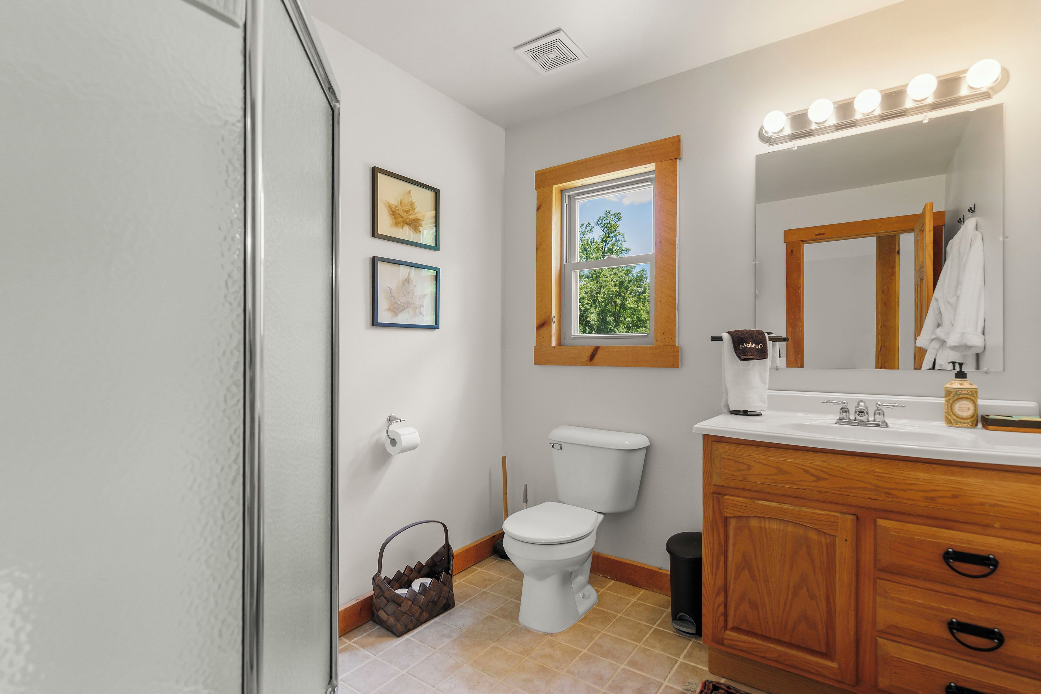 Une salle de bain comprenant des toilettes, un meuble-lavabo, un miroir et une fenêtre donnant sur des arbres.