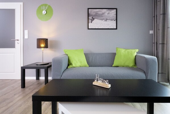 A modern living room with a grey sofa, two green cushions, a black coffee table, a small side table with a lamp, a framed picture on the wall, and a green clock.