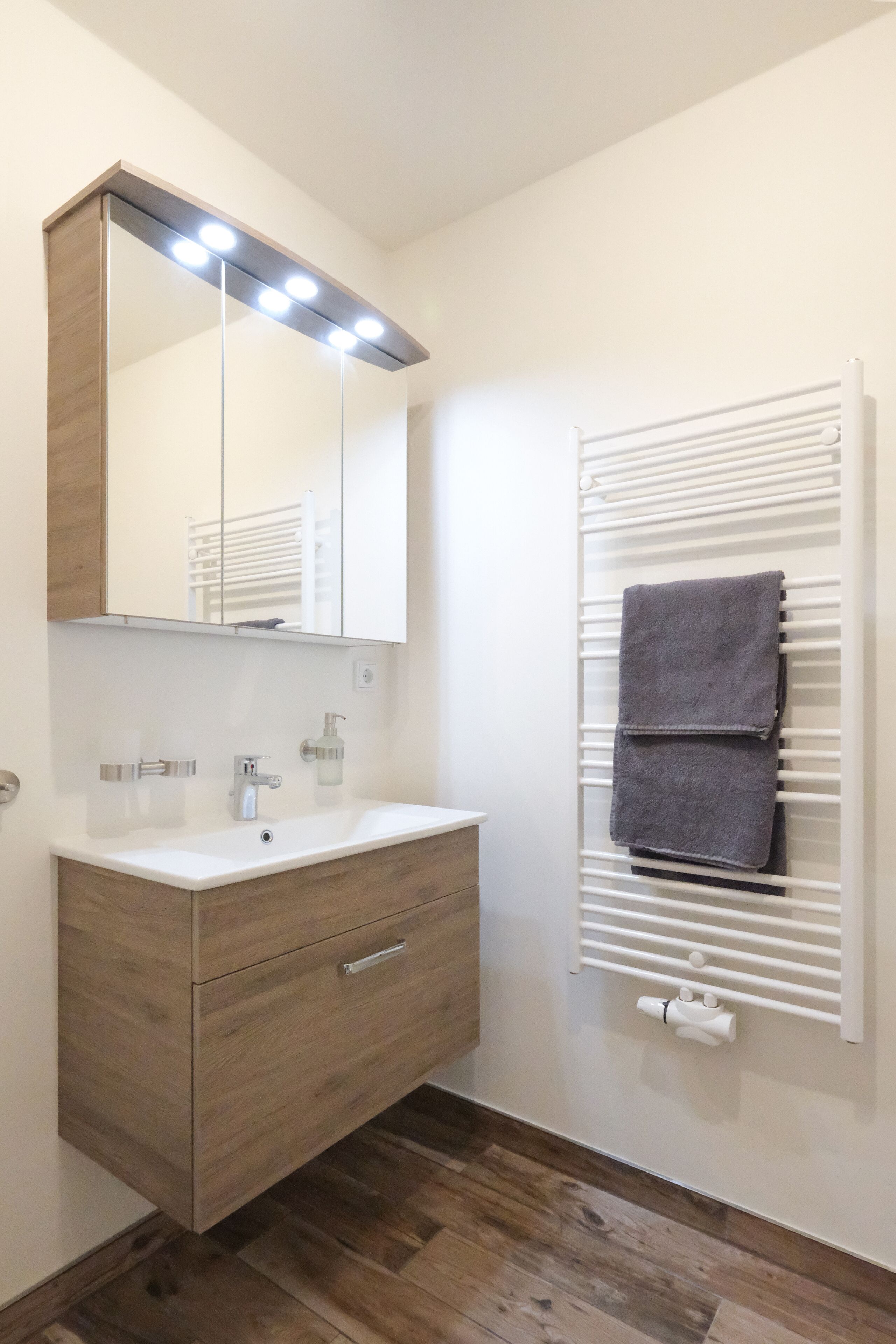 Un baño con tocador de madera, un espejo con luces, un perchero para toallas y un radiador blanco.