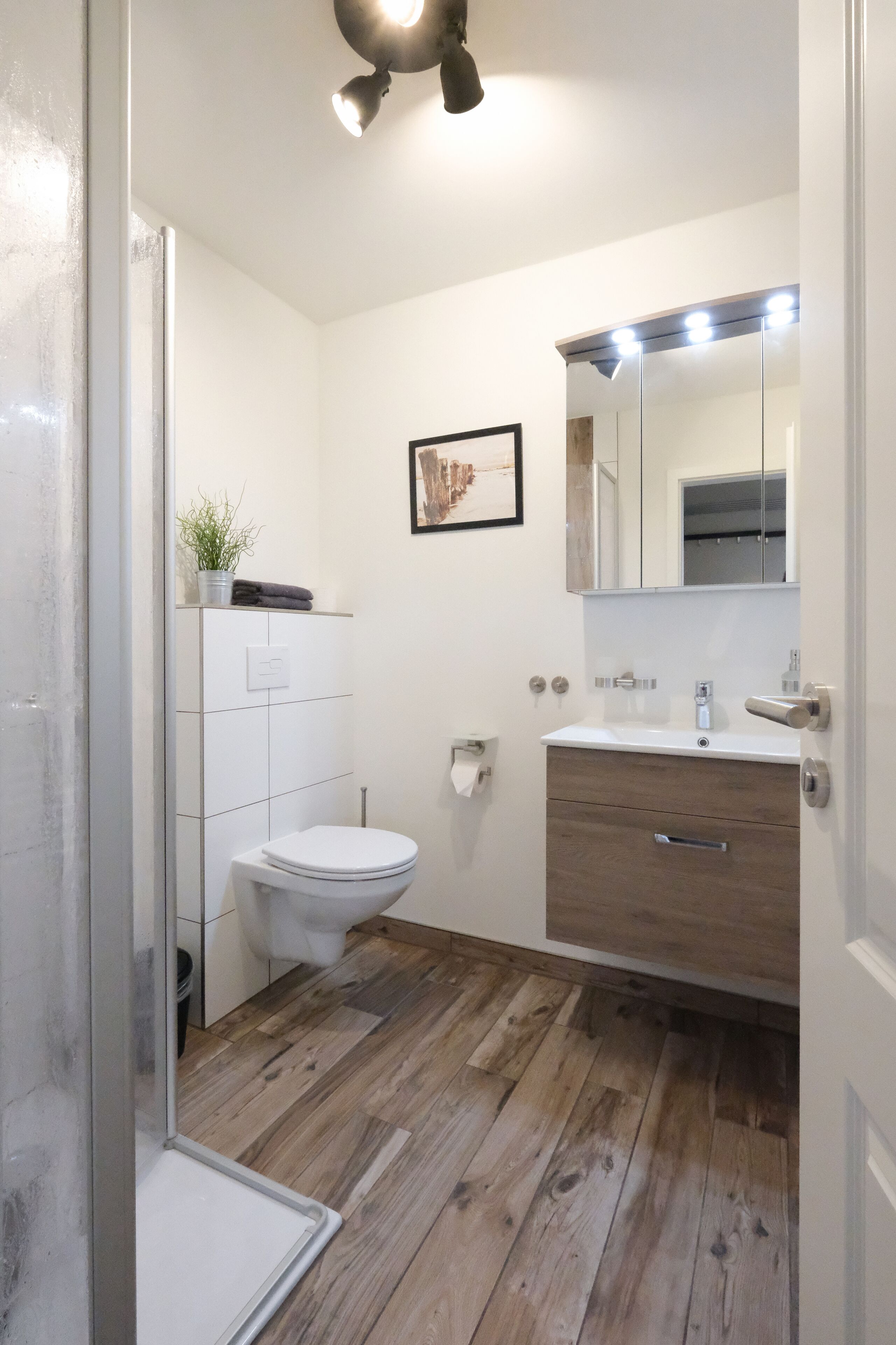 Ein modernes Badezimmer mit Holzboden, weißen Fliesen, Toilette, Waschbecken mit Spiegel und einer kleinen Pflanze an der Wand.