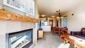 A cozy living room with a fireplace, a dining area with a wooden table and chairs, and a kitchen in the background.