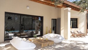 Un espacio exterior moderno con sofás blancos y una mesa de centro de madera, puertas de cristal grandes que dan a una piscina y una pérgola con plantas trepadoras.