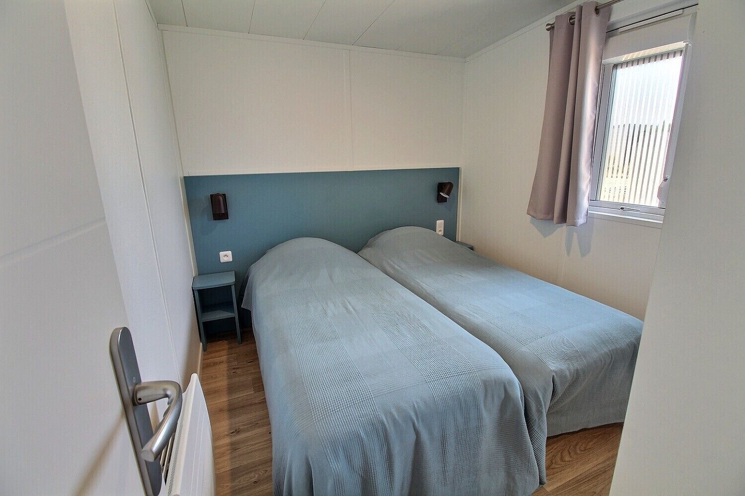 A small, single-room accommodation with two beds, a nightstand, a wall-mounted lamp, and a window with curtains.