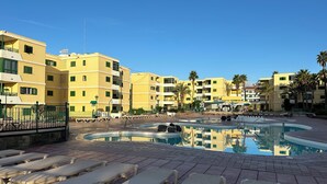 Uma área de piscina com espreguiçadeiras, céu claro e edifícios amarelos ao fundo.