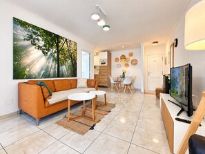 A modern living room with a sofa, coffee table, and TV. There is a dining area with chairs and a table. The room has a large painting on the wall and a kitchen in the background.