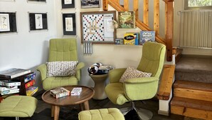 A cozy living room with green armchairs, a coffee table, and a bookshelf with board games. A wooden staircase with a railing is visible in the background. - Ashford cabin,Mt. Rainier view. Private river access, 17 acres, 5 mi to MRNP (Ashford)