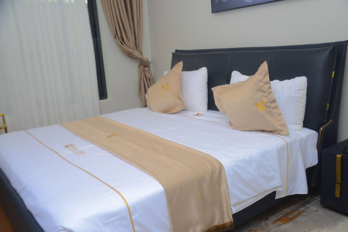Una cama bien hecha con una colcha blanca y beige, un cabecero oscuro y dos almohadas con un pequeño emblema.