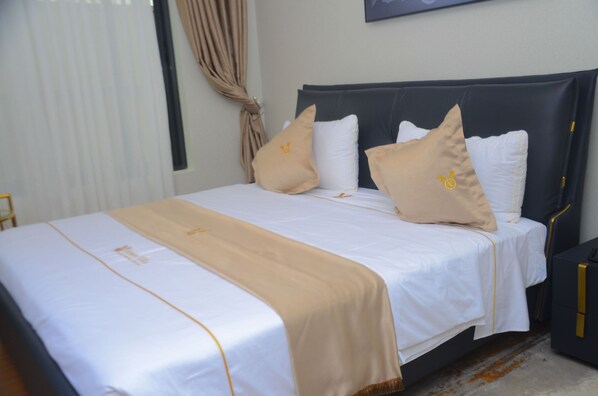 A neatly made bed with a white and beige bedspread, a dark headboard, and two pillows with a small emblem. - Victory Villa Apartment Hotel (Kigali)