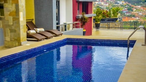 Ein Swimmingpool mit gefliesten Rändern, Liegestühlen und Blick auf eine Stadt im Hintergrund.
