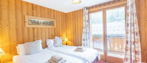 A wooden-paneled bedroom with two beds, a nightstand, a lamp, and a window with curtains.