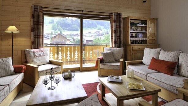 Una habitación acogedora con paredes de madera y un ventanal que ofrece vistas a un pueblo. Hay dos sofás, una mesa con velas y una estantería.