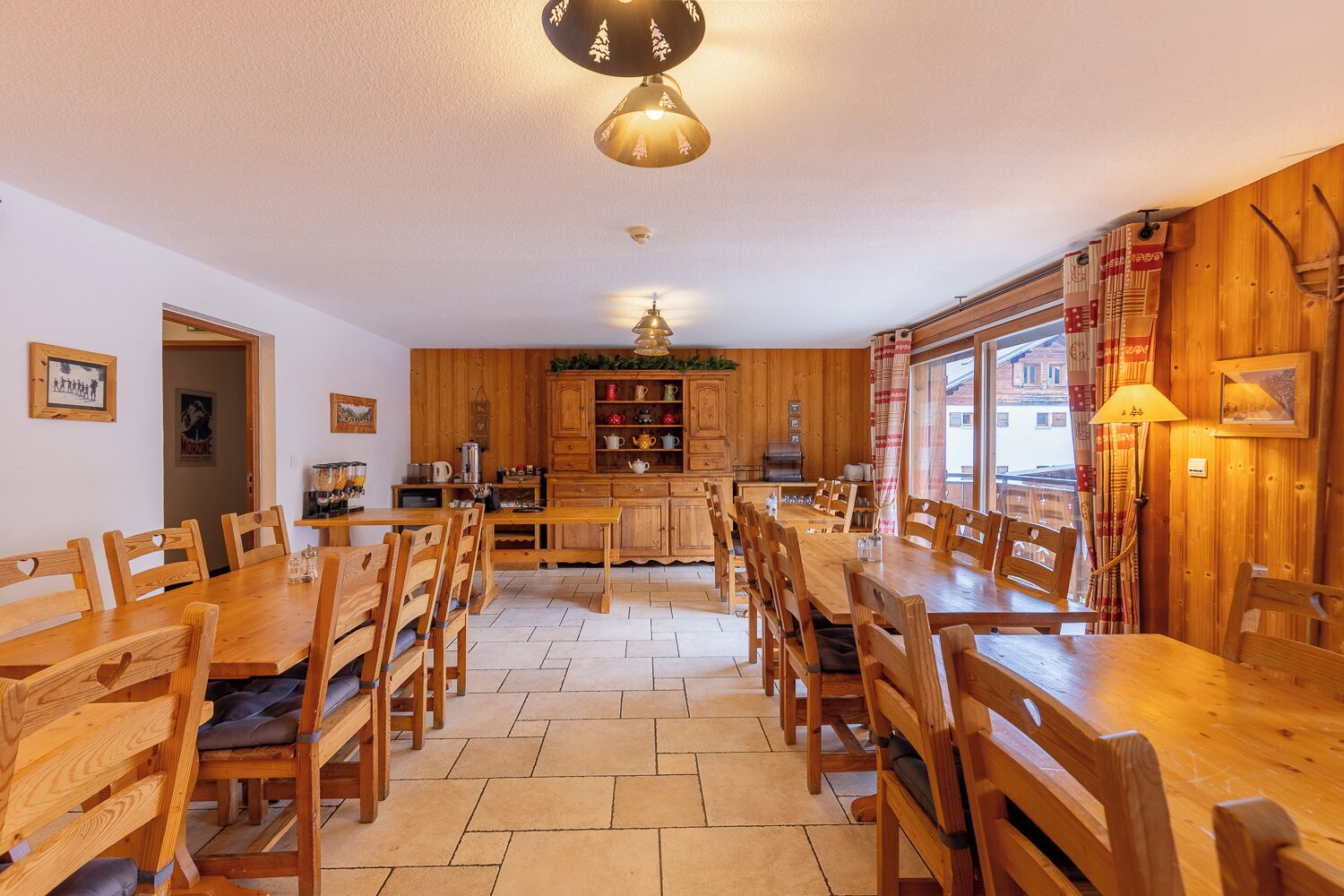 Un coin repas chaleureux avec des tables et des chaises en bois, une cuisine dotée de meubles en bois et une grande fenêtre agrémentée de rideaux.