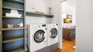A laundry room with two washing machines, shelves with folded towels, and a painting on the wall. - The Adaline #1 -  Luxury Hyde Park Apt Near Bayshore (Tampa)