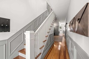 Un pasillo con piso de madera, paredes blancas y una escalera con barandilla blanca y escalones de madera.