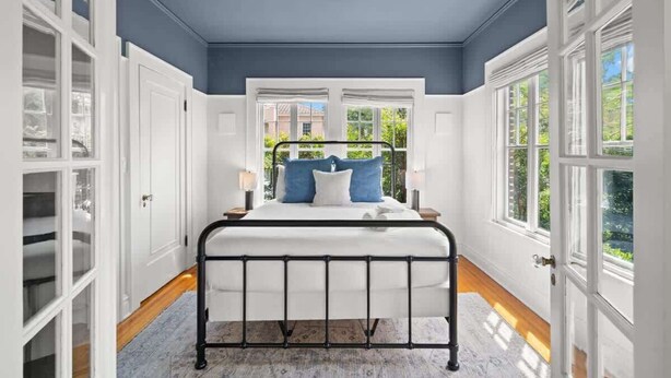 Ein Schlafzimmer mit Bett, weißen Wänden, blauer Decke und großen Fenstern.