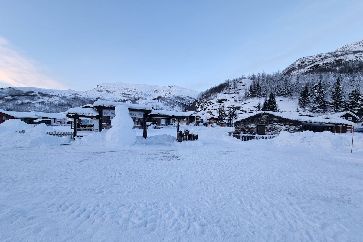 A snow-covered mountain village with rustic buildings, a wooden signboard, and a snowman.
