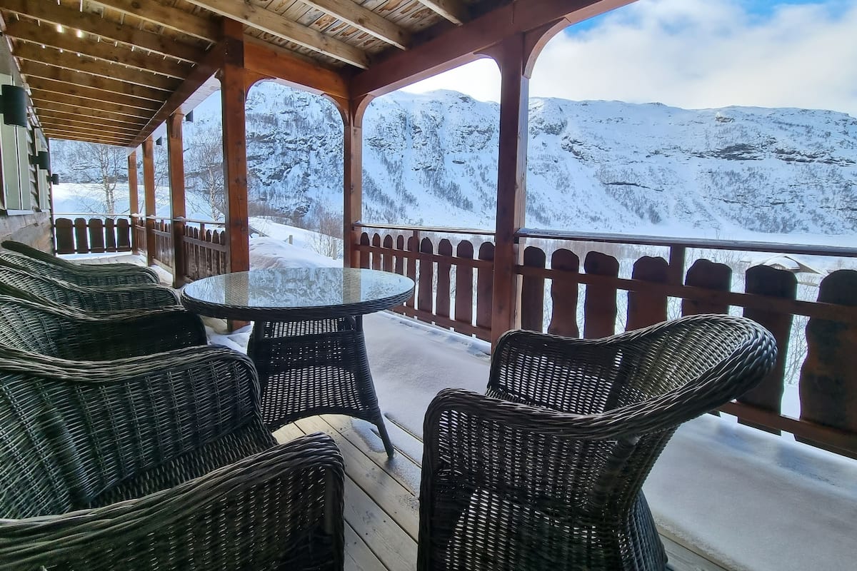 A covered outdoor seating area with wicker furniture and a glass table, overlooking a snowy mountain landscape.