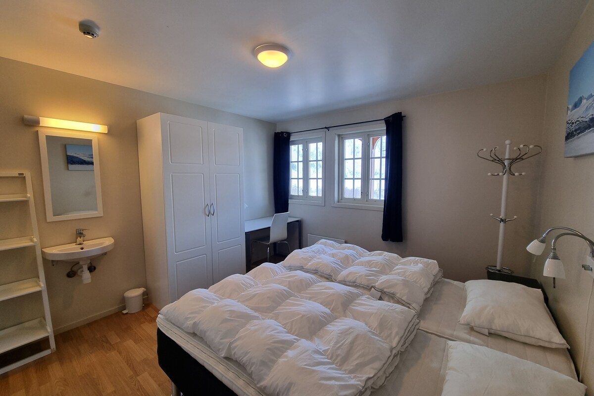 A bedroom with a bed, a bedside table, a lamp, a coat rack, a window, and a bathroom with a sink and toilet.