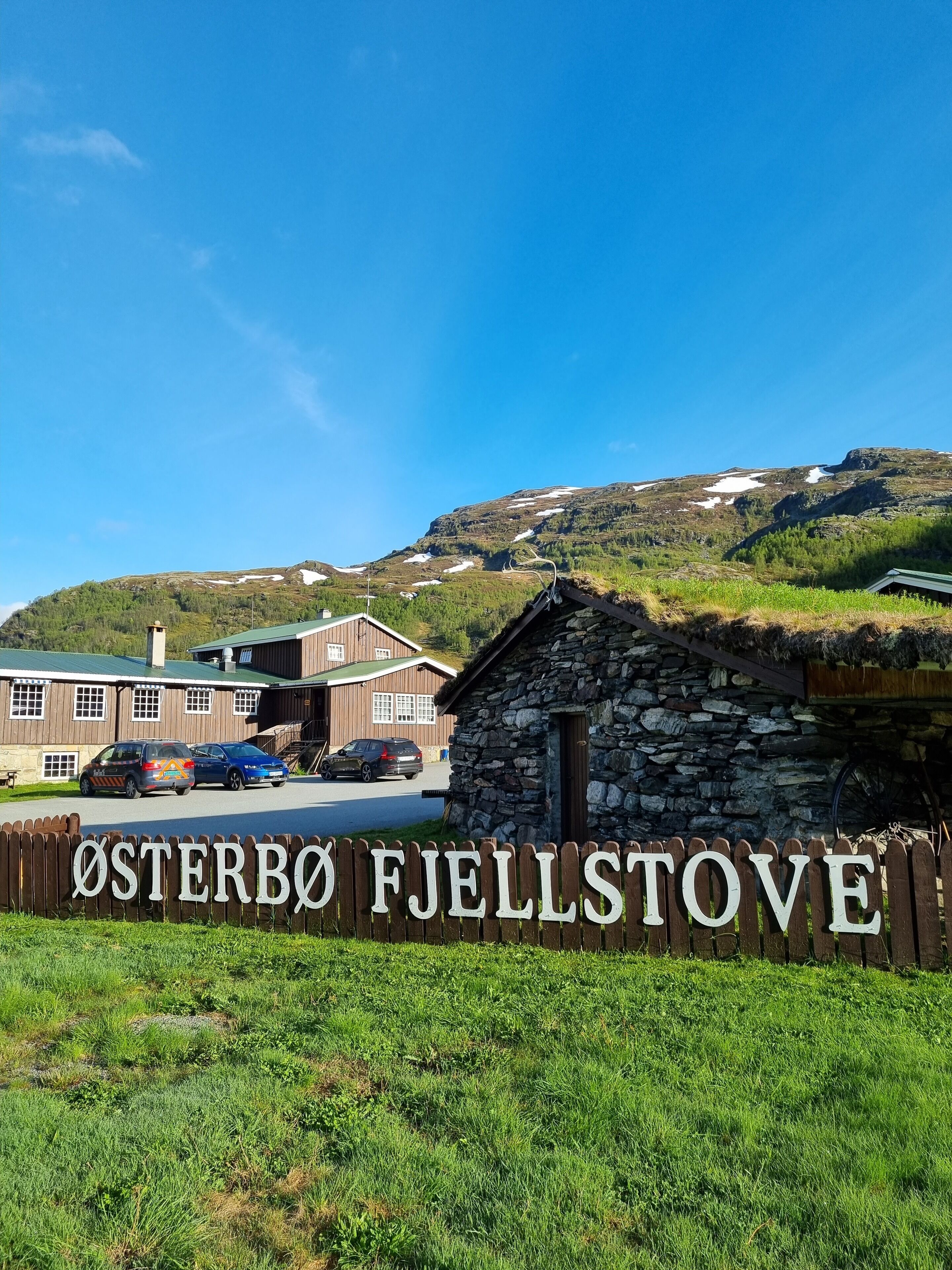 A wooden sign with the text "ØSTERBO Fjellstove" in front of a building with a green roof and a stone structure to the right.