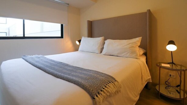 A neatly made bed with a patterned throw, a bedside lamp, and a window with blinds. - w* | Art Deco 1BR in Barranco (Barranco)