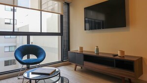 A modern living room with a large window, a blue chair, a round coffee table, a flat-screen TV mounted on the wall, and a wooden entertainment unit. - w* | Art Deco 1BR in Barranco (Barranco)