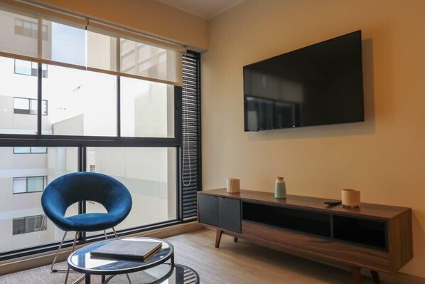 A modern living room with a large window, a blue chair, a round coffee table, a flat-screen TV mounted on the wall, and a wooden entertainment unit. - w* | Art Deco 1BR in Barranco (Barranco)