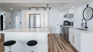 A modern kitchen with white cabinetry, a central island with stools, stainless steel appliances, and a hanging mirror. - Sandcastle 6A Completely Renovated Modern Condo On the Water Steps to the Beach (Dauphin Island)